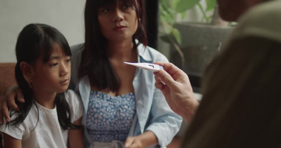 A mother checks a childs temperature with a thermometer while another adult observes in a plainly lit room