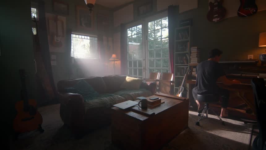 A person plays a piano in a room filled with musical instruments a couch and sunlight streaming through a window