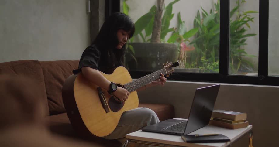 A young woman sitting on a sofa and playing the guitar while looking at a laptop in front of her