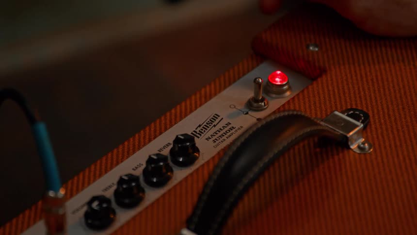 A hand reaches in to flip the power switch on an orangecolored benson nathan junior guitar amplifier a red light illuminates indicating the amplifier is now on
