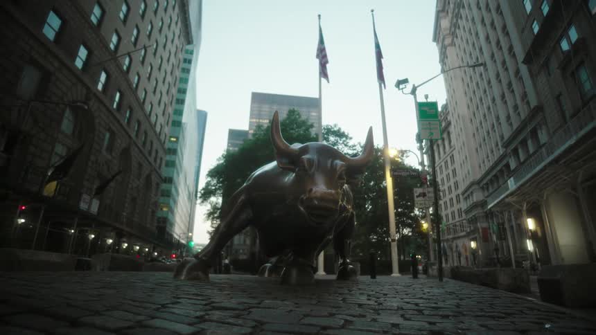A bronze bull statue stands on a cobblestone street flanked by tall buildings and flags in a dense urban setting