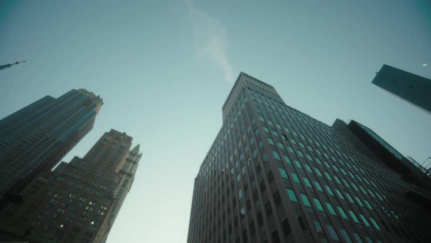 A towering group of modern city skyscrapers reaches towards a bright hazy sky as viewed from a low angle on a city street