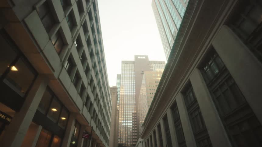 Several tall city buildings with numerous windows line a street creating a narrow view of a bright clear sky above
