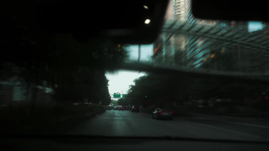 Car windshield driving through a city street with tall buildings and traffic at dusk