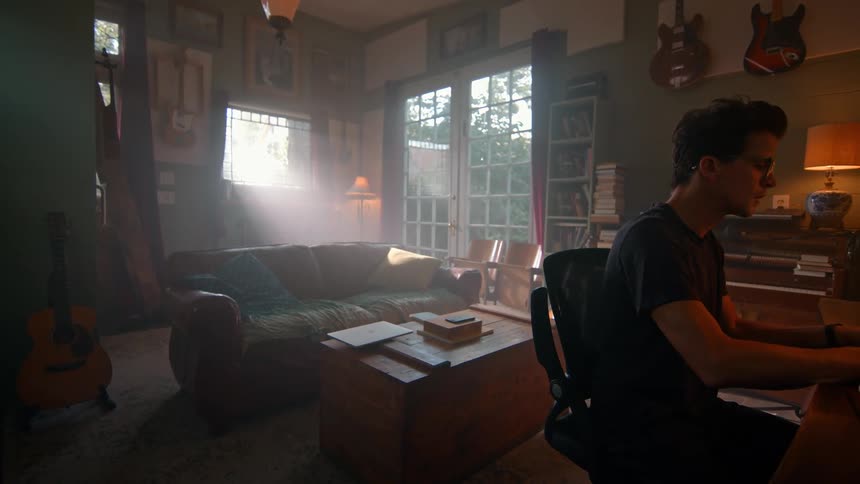 A person works at a desk in a sunlit room with green walls surrounded by guitars bookshelves and a brown leather couch