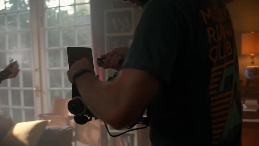 A musician plays guitar while a videographer films him in a softly lit room
