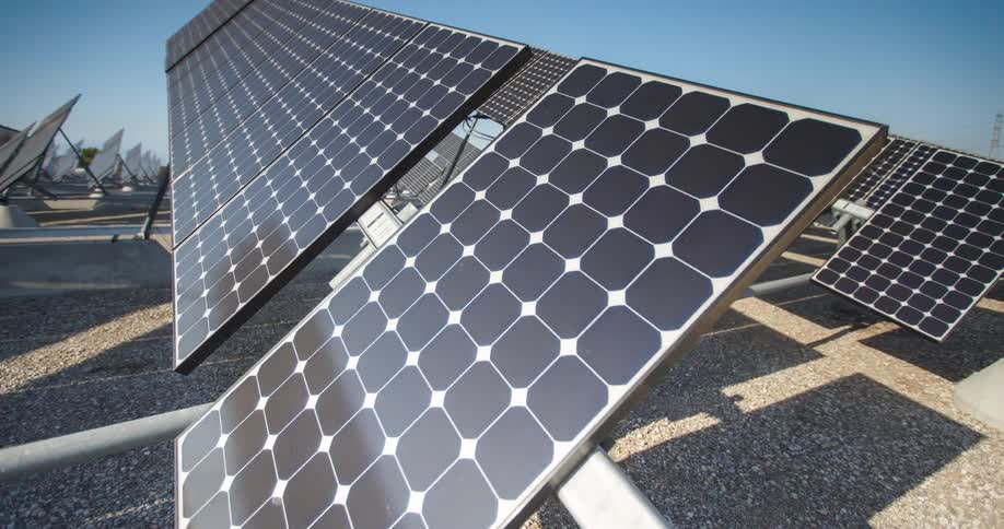 An array of dark solar panels with white grid patterns stands angled towards a bright blue sky