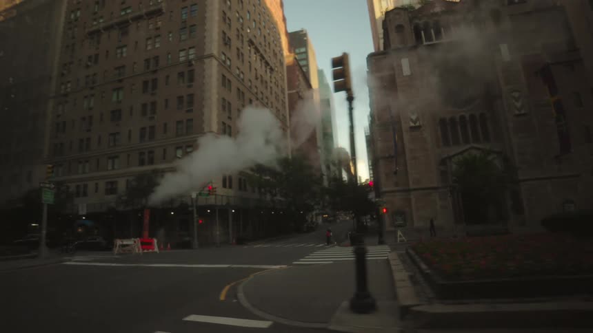 An intersection on a city street is lined with tall buildings with steam or smoke rising in the midground the traffic light at the intersection is red and the scene is set at sunset