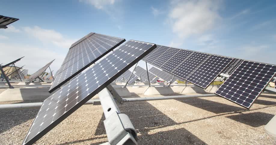 Angle shot of solar panels attached to metal frames tan gravel ground is beneth the panels abouve panels is a blus sky with scattered clouds