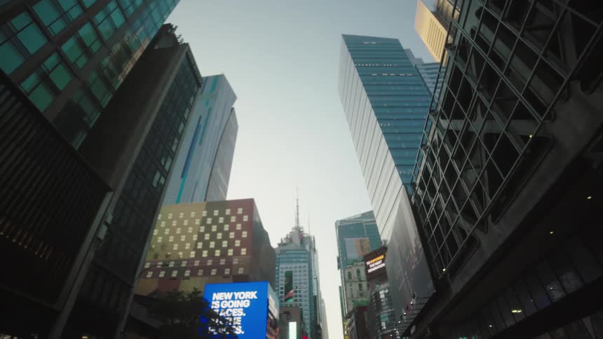 Multiple towering city buildings with many windows and brightly lit digital billboards rise towards a pale sky creating a dense urban landscape