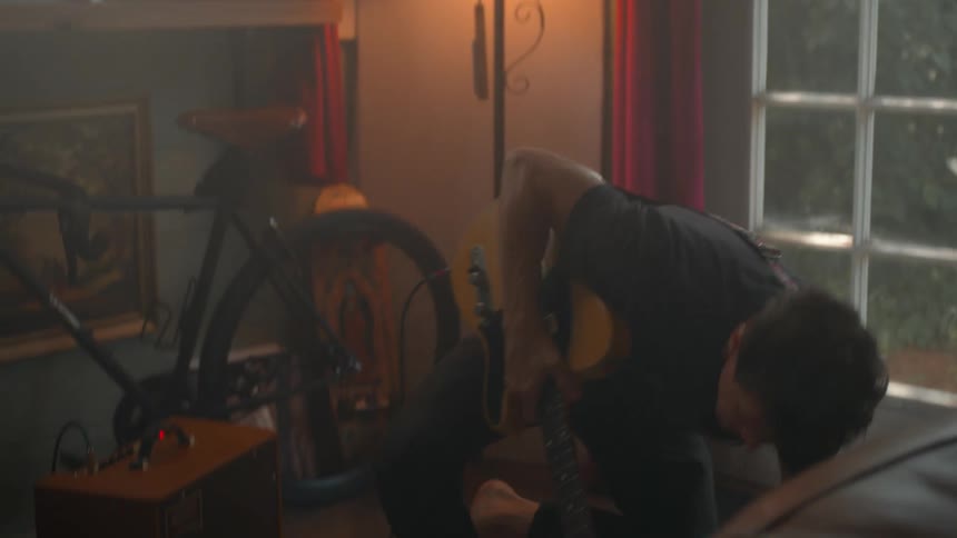 A cozy room corner holds a bicycle guitar and amplifier softly illuminated by a nearby lamp as a man plays guitar