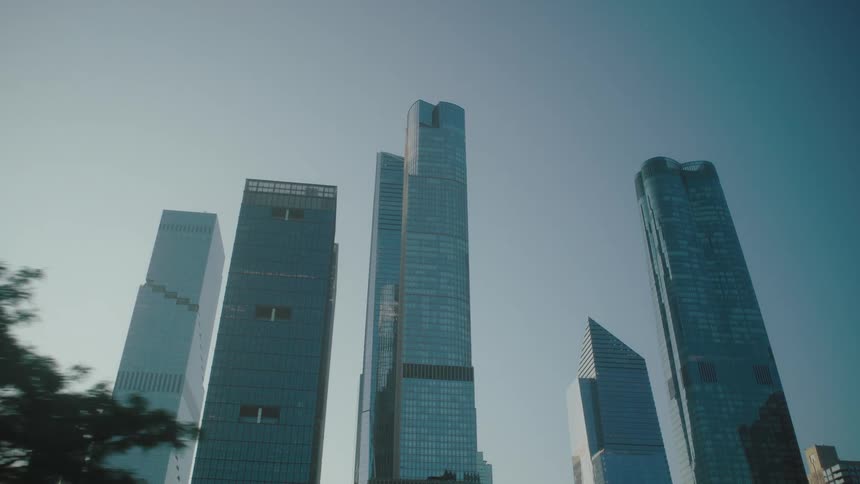 The video showcases tall modern skyscrapers against a clear sky suggesting a vibrant city setting as the camera pans lush green trees partially obscure the view of the buildings adding a touch of nature to the urban landscape