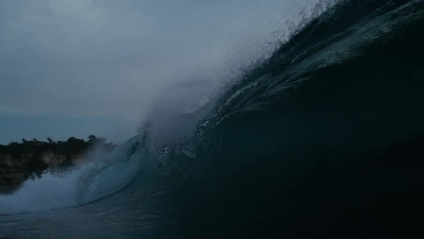 The video captures the raw power of a large ocean wave as it curls and breaks with white foam cascading down its face on the left side of the frame a rocky coastline with some green foliage is visible providing a rugged backdrop to the dynamic sea