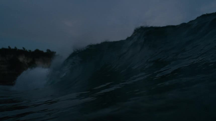 A large dark wave breaking under a cloudy sky with a rocky cliff in the background