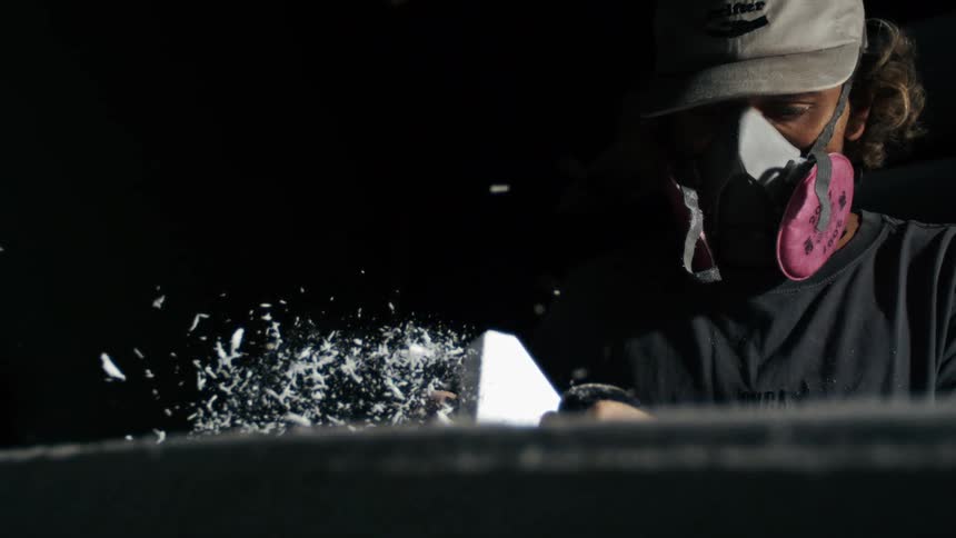 A person wearing a white baseball cap and a grey respirator with pink filters is intensely focused on their work they are seen from the chest up with their head slightly tilted to the right the person is using a tool to shave white material