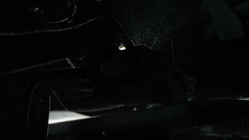 A person wearing a hat and respirator mask works with a handheld tool sending a spray of particles into the air under a bright overhead light in a dark workshop
