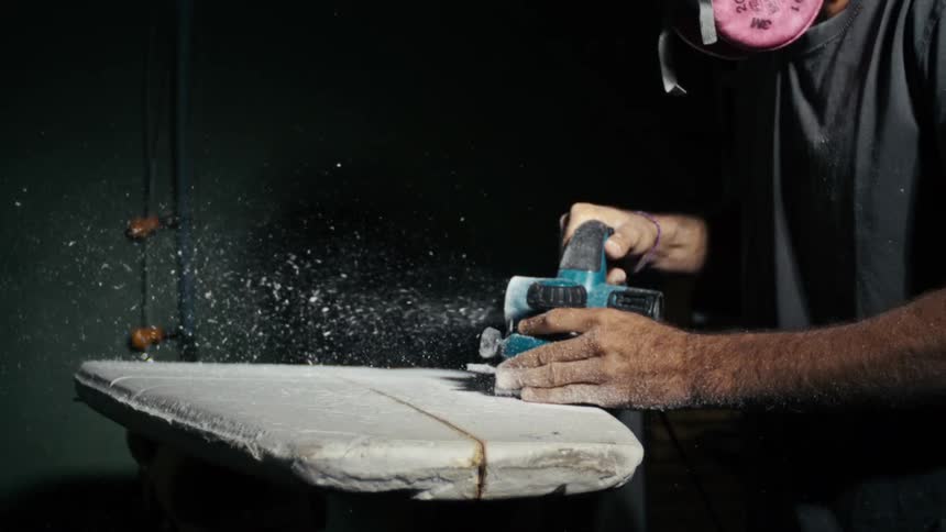 Man uses an electric sander to shape a surfboard in a dark room