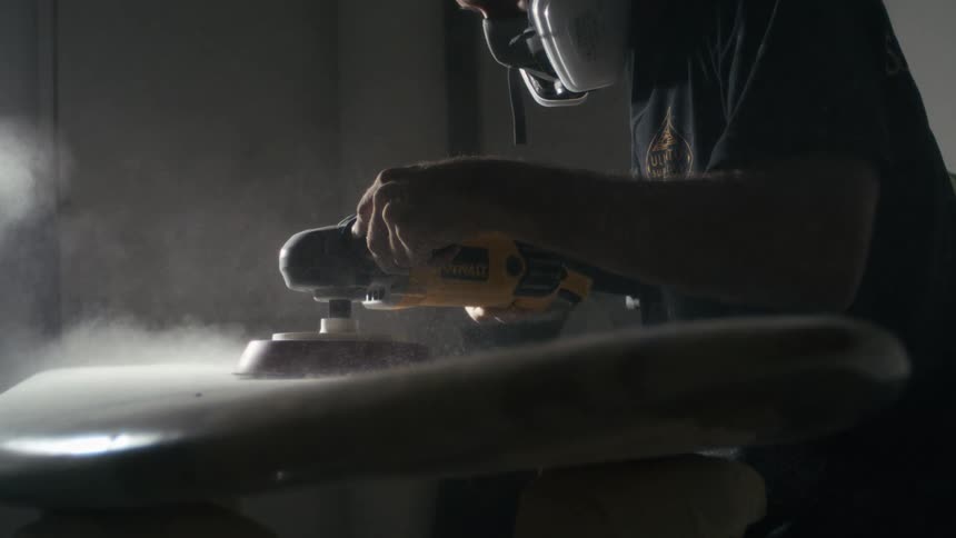 A craftsman wearing a respirator shapes a surfboard in a dimly lit workshop filled with dust and focus