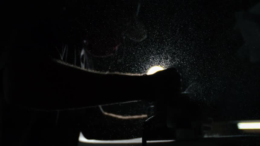 A person wearing a protective face shield grinds metal under bright workshop lighting sending dust and debris outward
