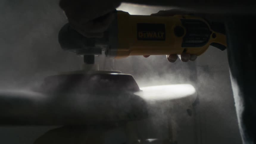 A carpenter is using a sander tool to sand a surfboard inside a workshop