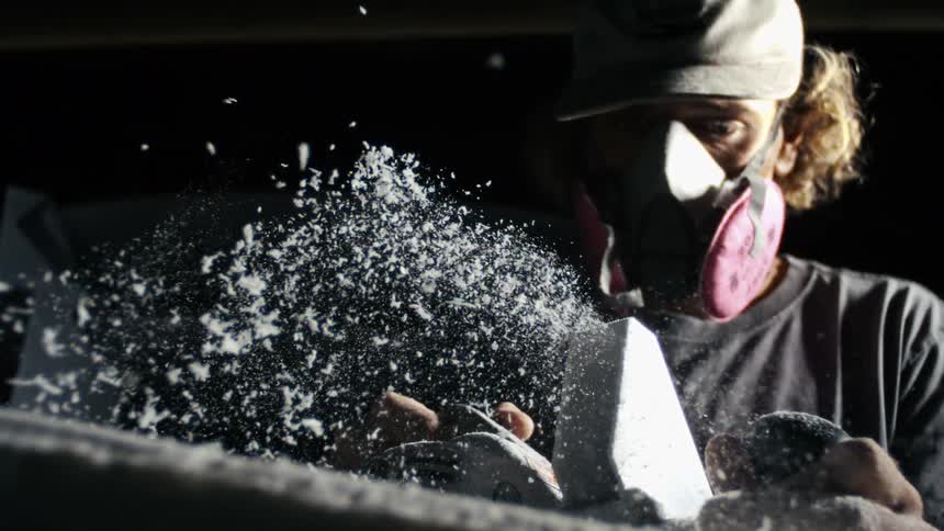 Man wearing a protective breathing mask uses a power tool to shave a pale surface filling the area with shavings