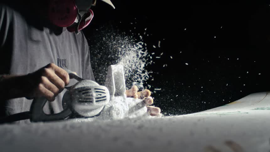 A craftsman shapes a surfboard with a power tool sending foam particles flying through the air