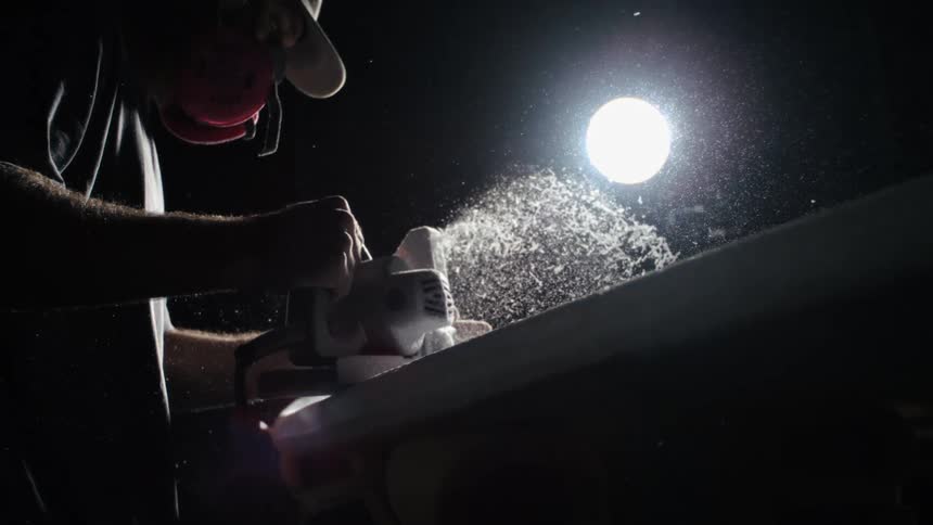 A person wearing a respirator and cap uses a planer on a piece of material creating a cloud of sawdust illuminated by a bright light