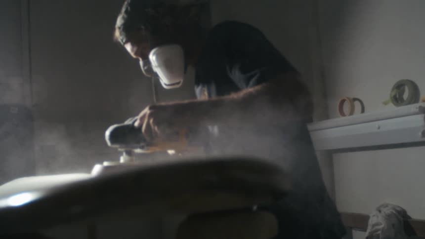 Man wearing a mask and using a polishing tool on a surfboard