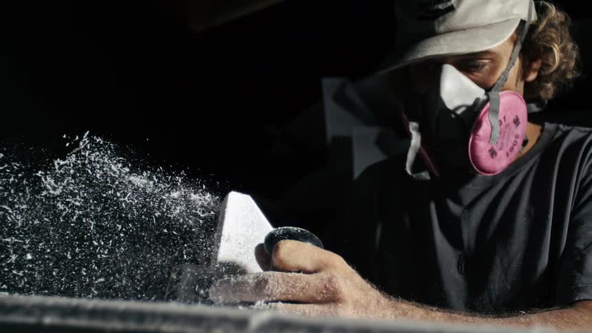 Man wearing a cap and mask using an electric sander to make a surfboard