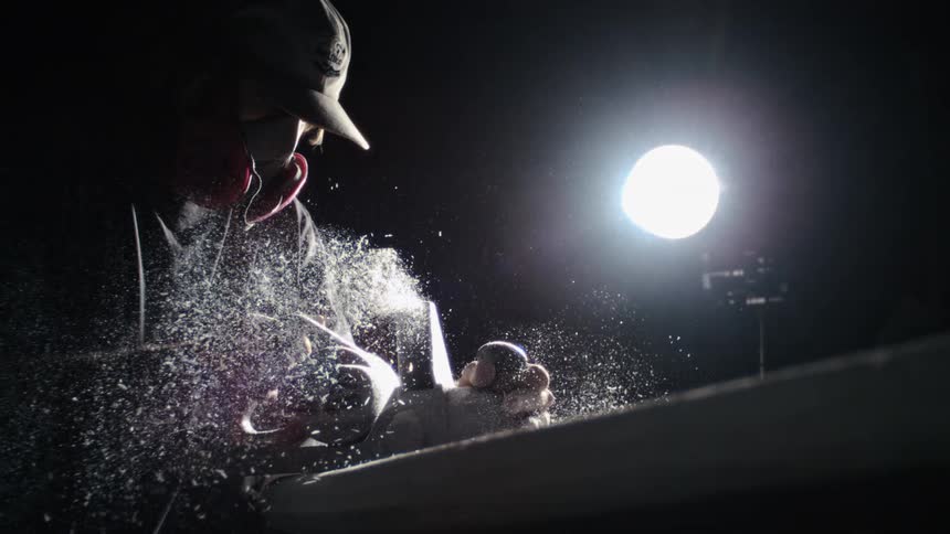A person wearing a hat and respirator mask works with a handheld tool sending a spray of particles into the air under a bright overhead light in a dark workshop