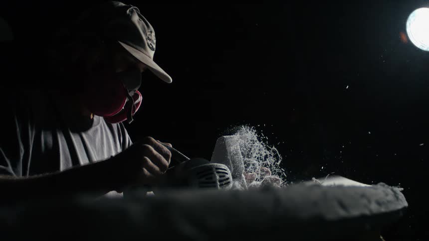 Man in cap and mask with a machine shaving a surfboard