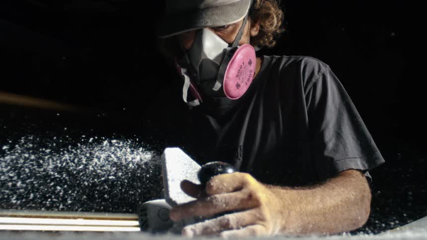 A person wearing a respirator mask sands a surface in a dusty room lit by a nearby window