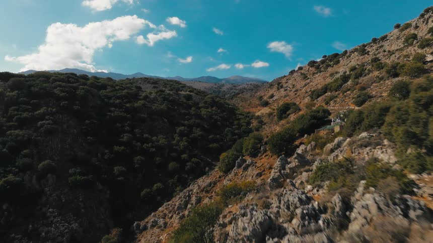 A person hikes across rocky terrain surrounded by forested mountains under a blue sky