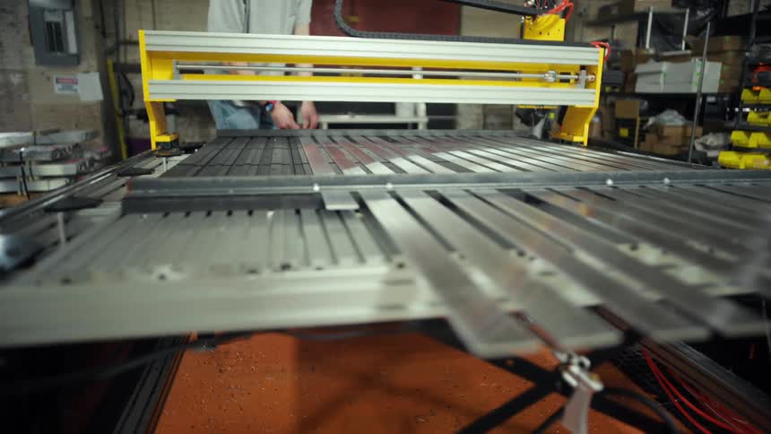 The video displays an industrial machine possibly a cnc router with a yellow gantry moving across a metalslatted bed a workers lower body is visible in the background of the workshop