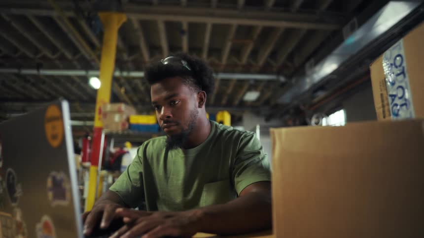 A person works on a stickercovered laptop at a wooden table in an industrial space filled with cardboard boxes and tools