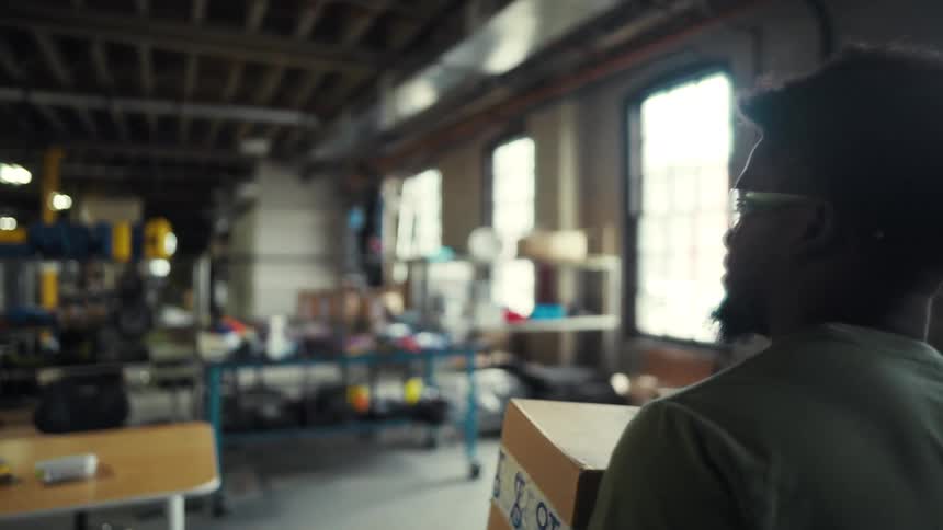 A young man walks into a distribution warehouse carrying shipping boxes and places them on a desk
