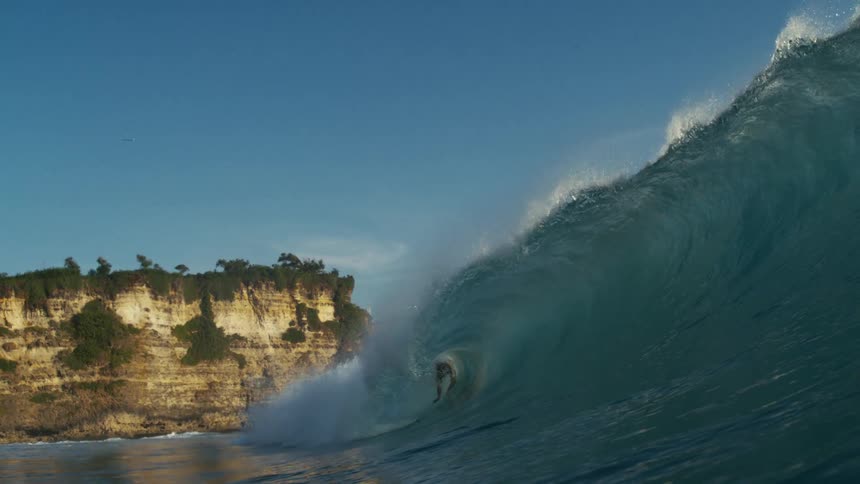 A surfer rides a large wave near a rocky coastline under a clear blue sky