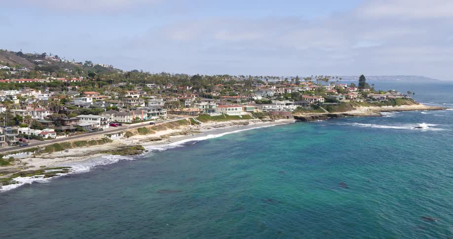 Coastal homes stretch along a rocky shoreline with turquoise waters gently meeting the sandy beach under a partly cloudy sky