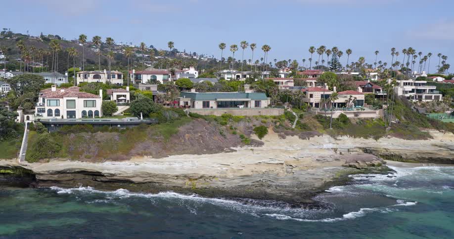 The video shows an elevated view of a luxurious coastal neighborhood where numerous multistory houses with redtiled roofs are perched atop a cliff overlooking the ocean palm trees dot the landscape adding to the picturesque scenery