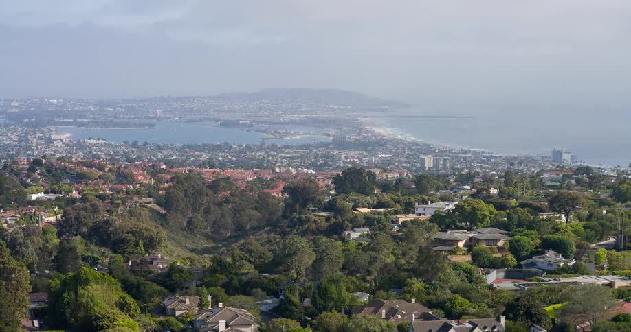 The video provides an expansive view of a sprawling city nestled beside a bay and the vast ocean residential areas with numerous houses are visible amongst lush green hills and trees creating a scenic urban landscape