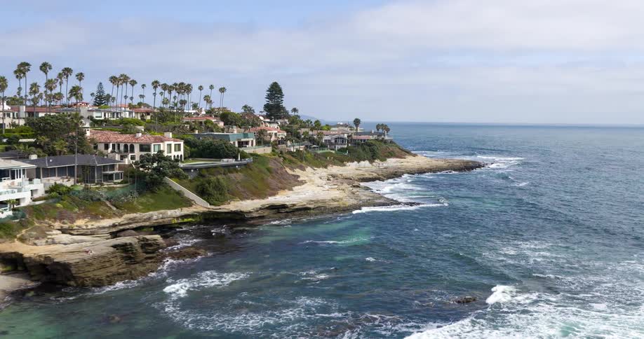 A coastal town with a series of residences sits along the shore as ocean waves hit the coastline at midday