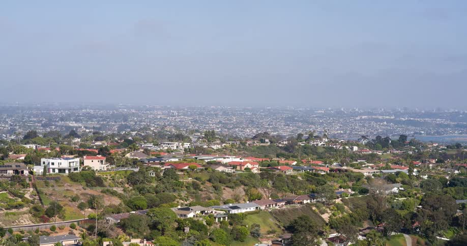 A winding road curves through a hilly residential area with redroofed houses and tall antennas atop the peak under a clear sky