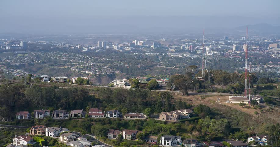 Suburban houses with red roofs sit at the edge of a wooded hill topped with communication towers with a sprawling city skyline visible in the distance