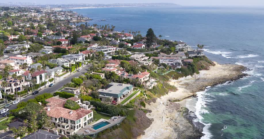 A coastal town with a series of suburban buildings sits by the ocean with waves hitting the shore at midday