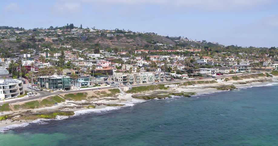 The video offers an elevated perspective of a winding coastal road with a cliffside adorned by lush greenery and palm trees on one side and the blue ocean with gentle waves on the other elegant multistory houses are nestled on the hillside