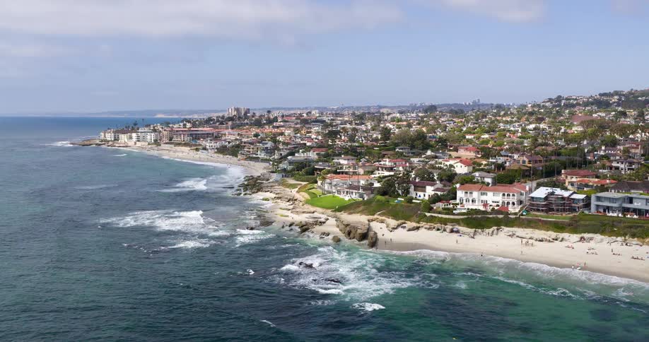 Aerial view of an urban waterfront with upscale beachfront homes and waves crashing against the rocks on a sunny beach