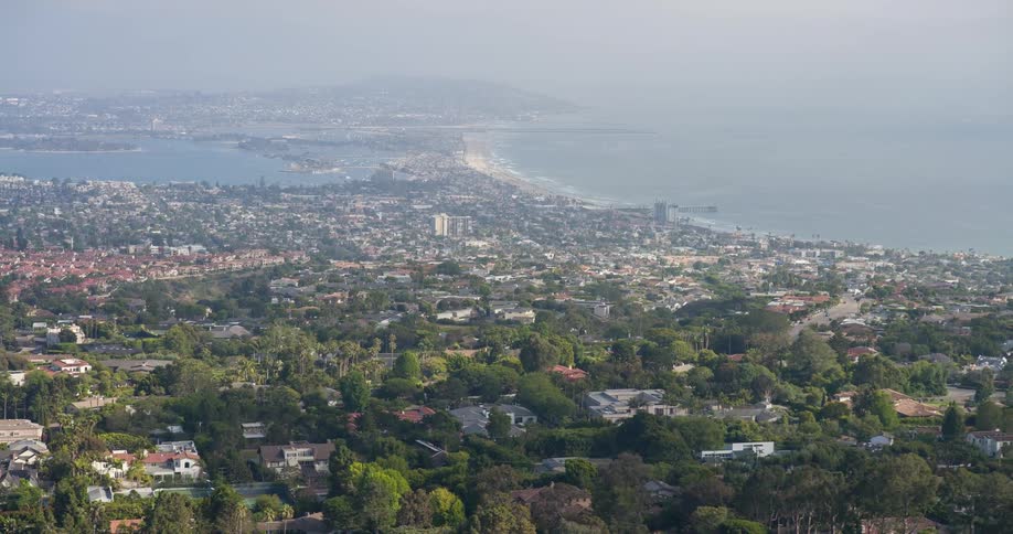 The video presents a highangle view of a vast urban landscape featuring numerous buildings and homes nestled among lush green trees in the background a large body of water possibly an ocean or bay stretches to the horizon under a hazy sky