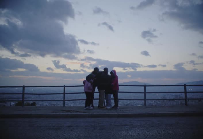 A family of five is standing on the side of the road beside a rail fence group hugging while wearing jackets on a cloudy evening