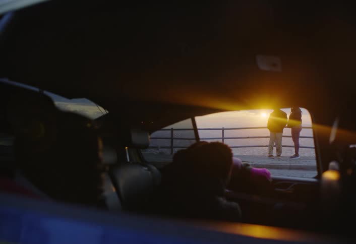 Two kids sit in a car and watch two adults converse on a sidewalk on a bridge