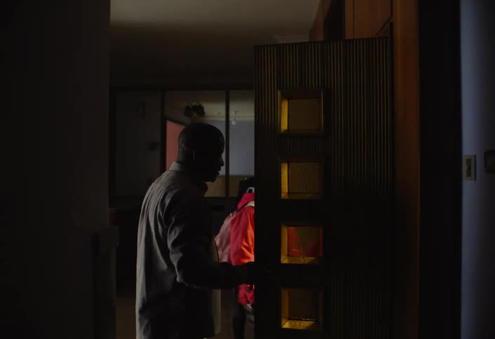 A father opens a wooden door with square glass panels as his family walks past him through a dimly lit room leaving home